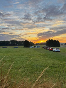 Mehrere Autos fahren in einer Reihe über ein Feld in Richtung Campingplatz. Im Hintergrund geht die Sonne am Horizont auf.