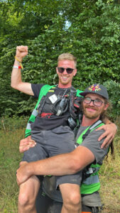 Ein Spektralwerk-Mitarbeiter trägt lachend einen Kollegen auf dem Arm. Beide schauen in die Kamera, tragen Warnwesten und Arbeitsequipment wie Walkie-Talkies.