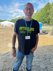 Ein Spektralwerk-Mitarbeiter im Nature-One-T-Shirt steht mit Mitarbeiterausweis um den Hals auf dem Festivalgelände und lächelt in die Kamera.
