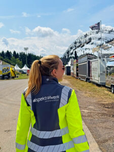 Eine Spektralwerk-Mitarbeiterin geht über das Festivalgelände und blickt über ihre rechte Schulter. Auf der Rückseite ihrer Warnweste ist das Spektralwerk-Logo zu sehen.