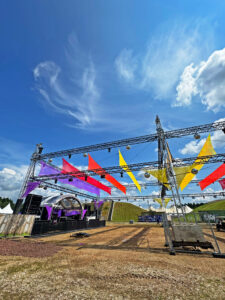 Eine Bühne auf der Nature One im Aufbau. An den Traversen vor der Bühne hängen bunte Stoffe in Regenbogenfarben. Der Himmel darüber ist strahlend blau.