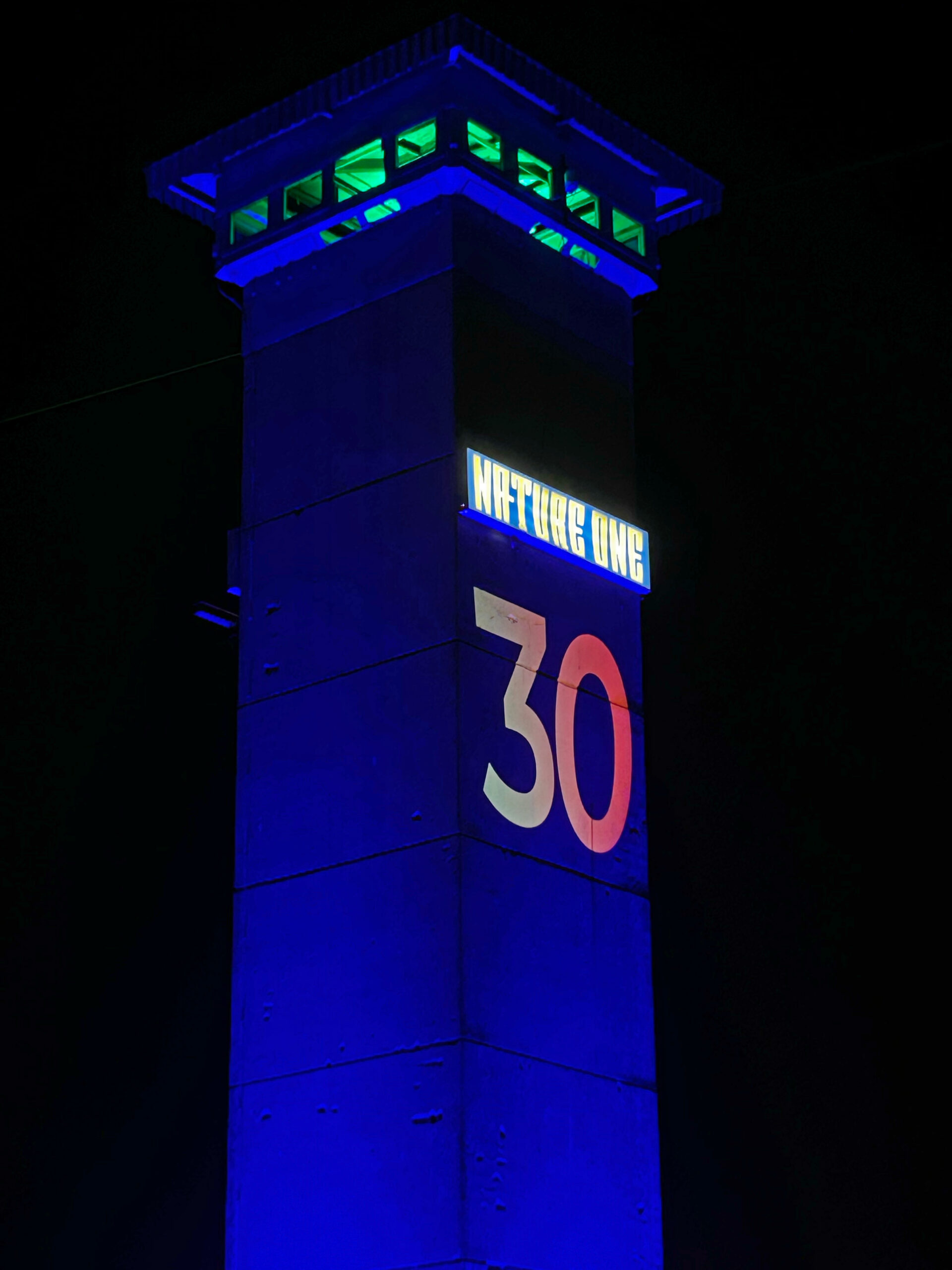 Ein blau beleuchteter Turm erhebt sich in den dunklen Nachthimmel. Auf seiner Vorderseite leuchten die Zahl ‚30‘ und ein Schild mit der Aufschrift ‚Nature One‘.